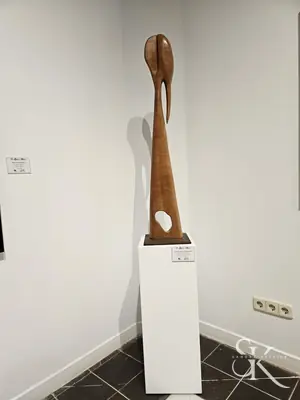 Escultura alta de madera con aberturas, sobre una peana blanca en una exposición
