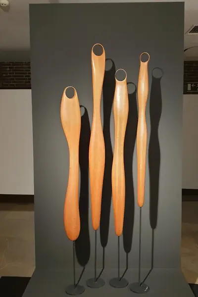 Tres esculturas altas de madera anaranjada, de formas onduladas, sobre pies negros en una galería