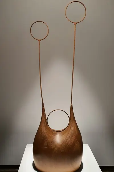 Escultura de madera oscura con una abertura y dos aros en lo alto, sobre peana blanca