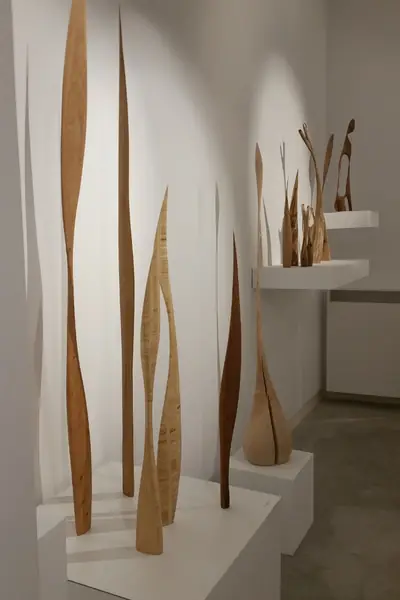 Tres piezas alargadas de madera clara, verticales, expuestas sobre peanas blancas en una galería