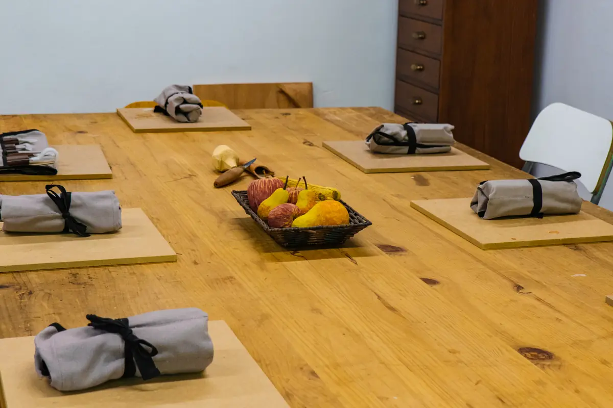Mesa larga de trabajo preparada para una clase, con los rollos de gubias y una bandeja de frutas talladas