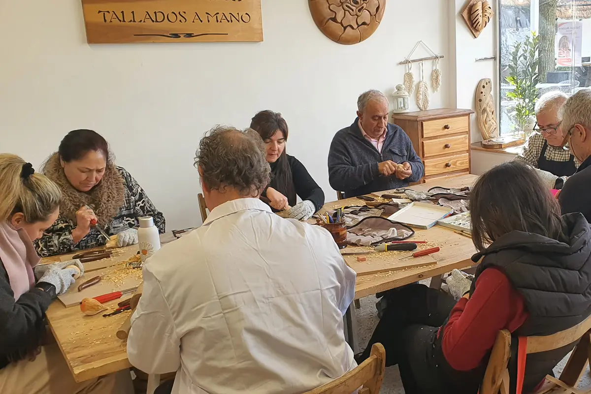 Clase completa alrededor de la mesa del taller, bajo el cartel tallado «Tallados a mano», cada alumno con su pieza