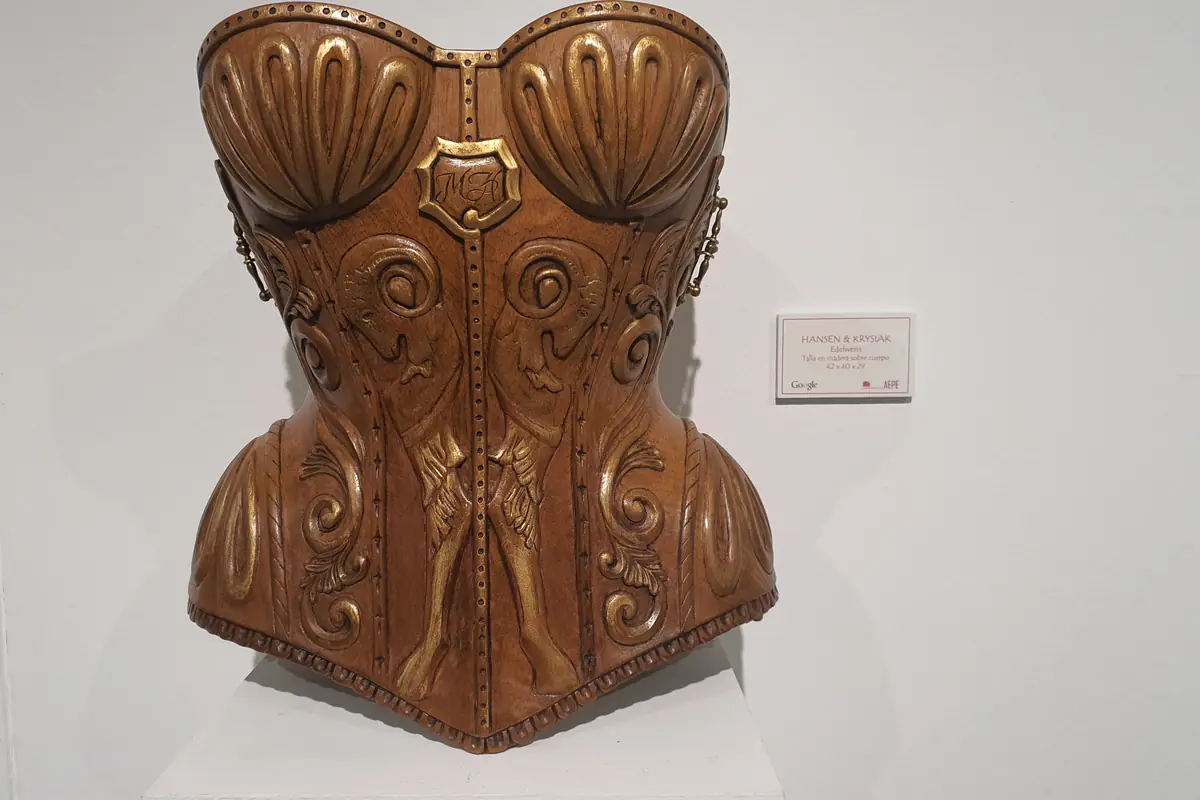Corsé tallado en madera sobre una peana blanca en la sala de exposición, con el relieve de los pliegues y el cierre marcados a gubia