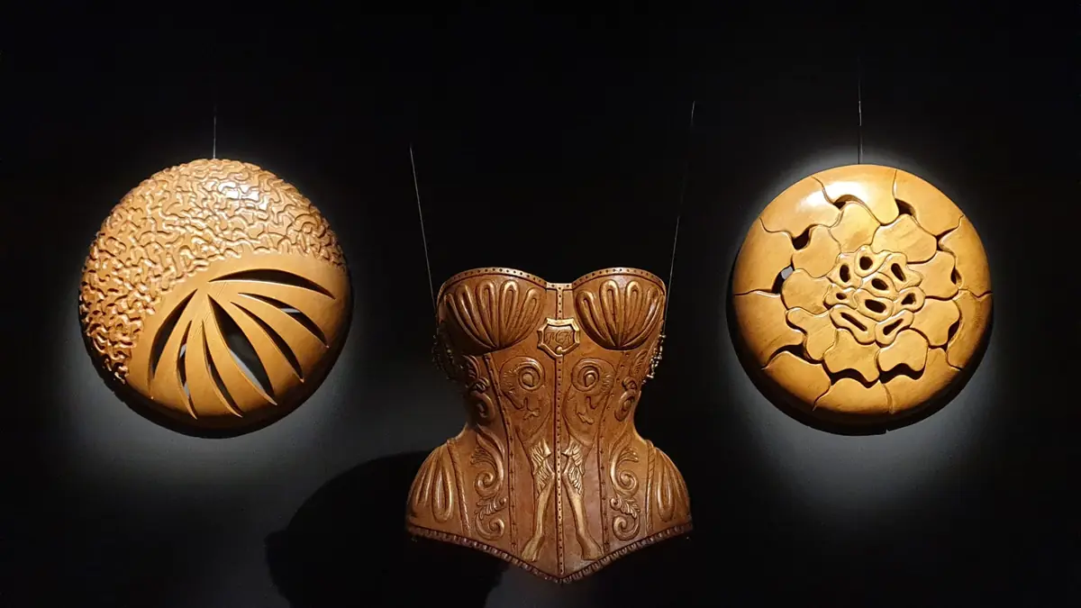 Tres piezas talladas en madera iluminadas sobre la pared negra de una galería: un corsé entre dos relieves circulares