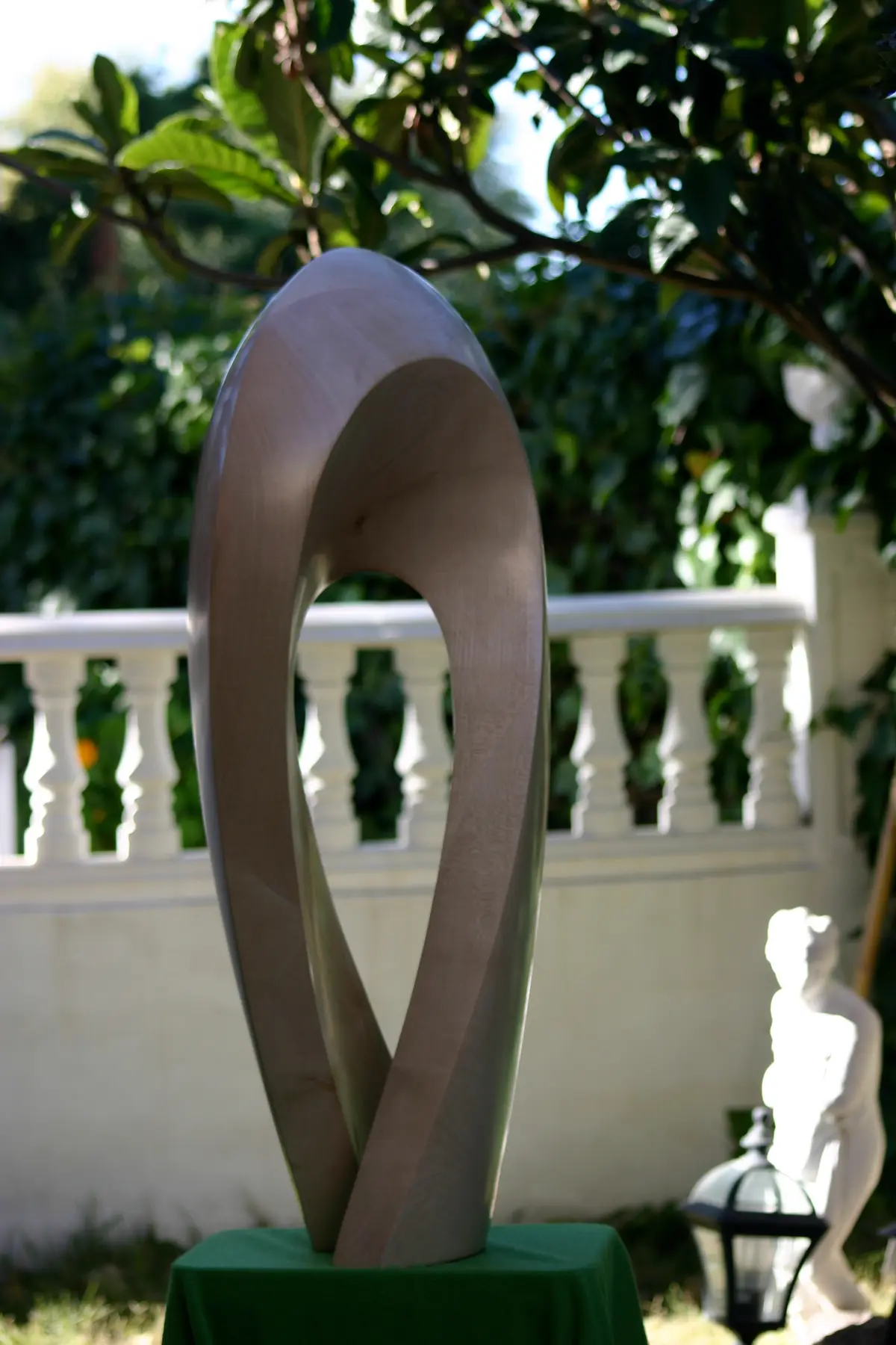 Escultura vertical de madera clara con forma de lazo cerrado que se retuerce sobre sí mismo, fotografiada en un jardín